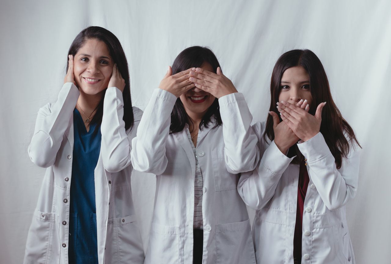 Three women scientists in lab coats posing playfully, symbolizing see no evil, hear no evil, speak no evil.